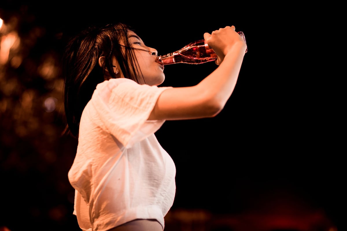 A woman in a white top drinks from a bottle outside at night, enjoying the refreshing beverage.