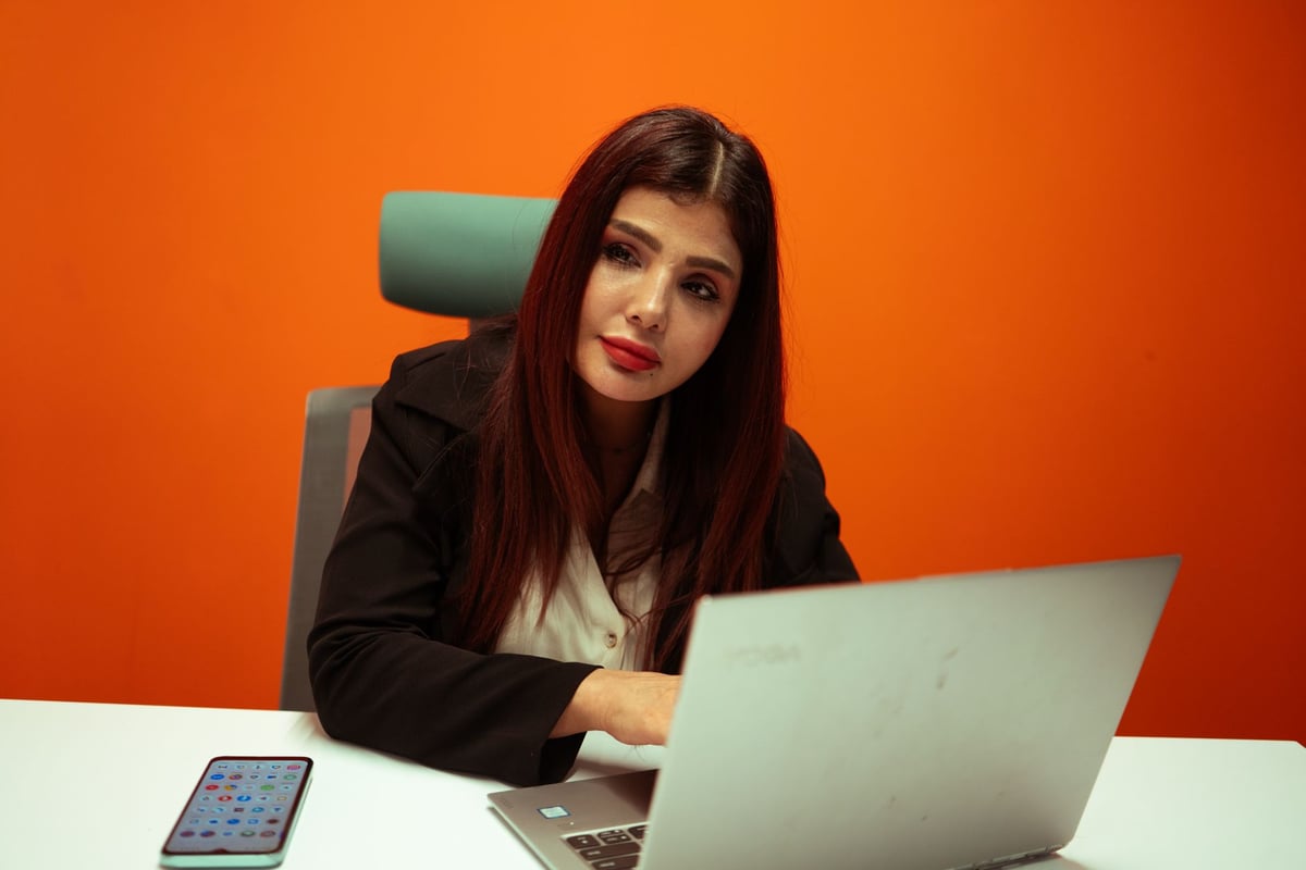 Focused businesswoman working on a laptop at a modern office desk with vibrant orange background.