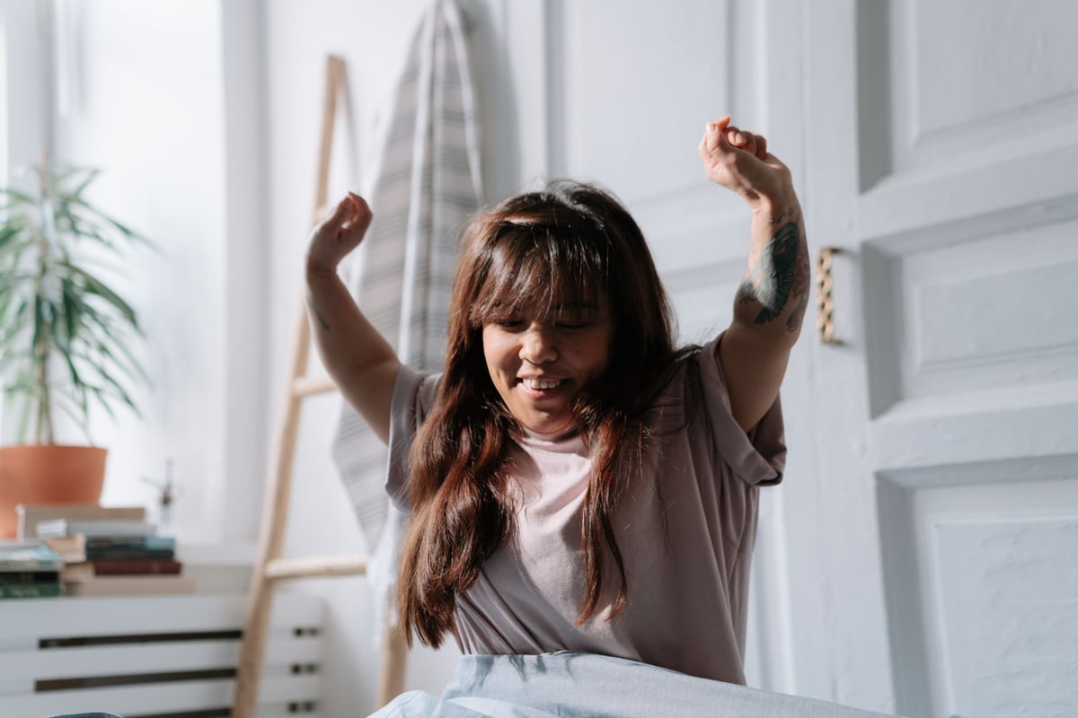 A relaxed Asian woman wakes up and stretches in a sunlit bedroom, enjoying the morning light.