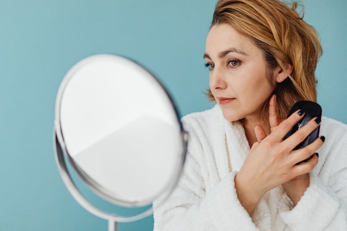 A woman brushes her hair in front of a mirror with a pastel blue background.
