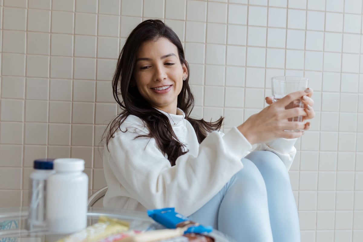Smiling woman holding a glass while seated, with pill bottles on the table.