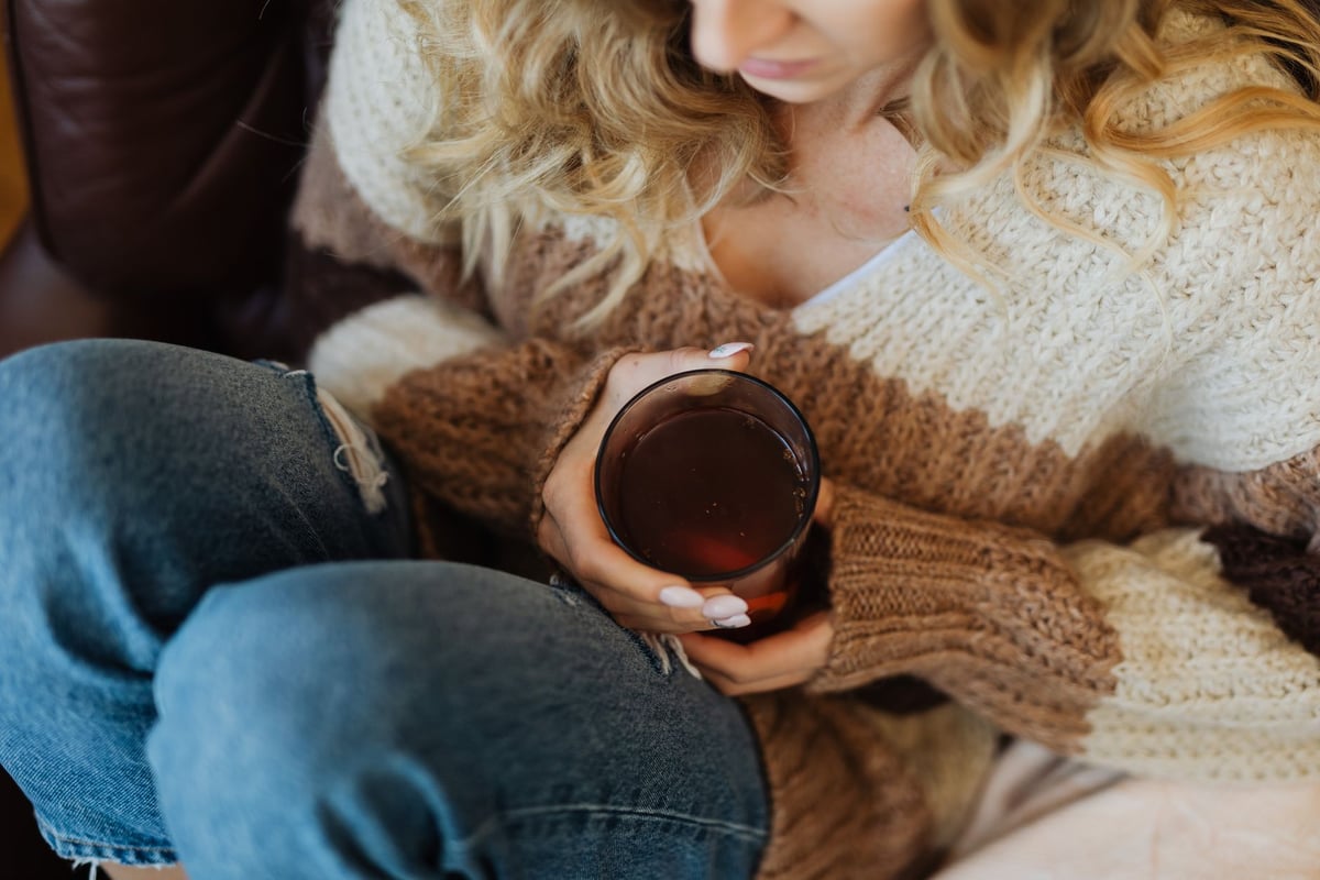 A woman in a cozy sweater and jeans holds a glass of tea, creating a warm indoor moment.