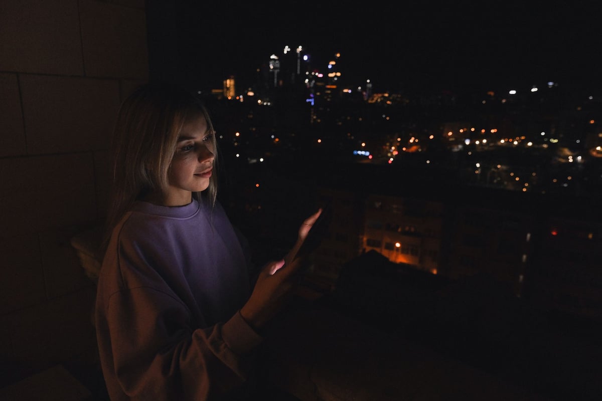 A woman on a balcony at night, illuminated by her smartphone, overlooking a cityscape.